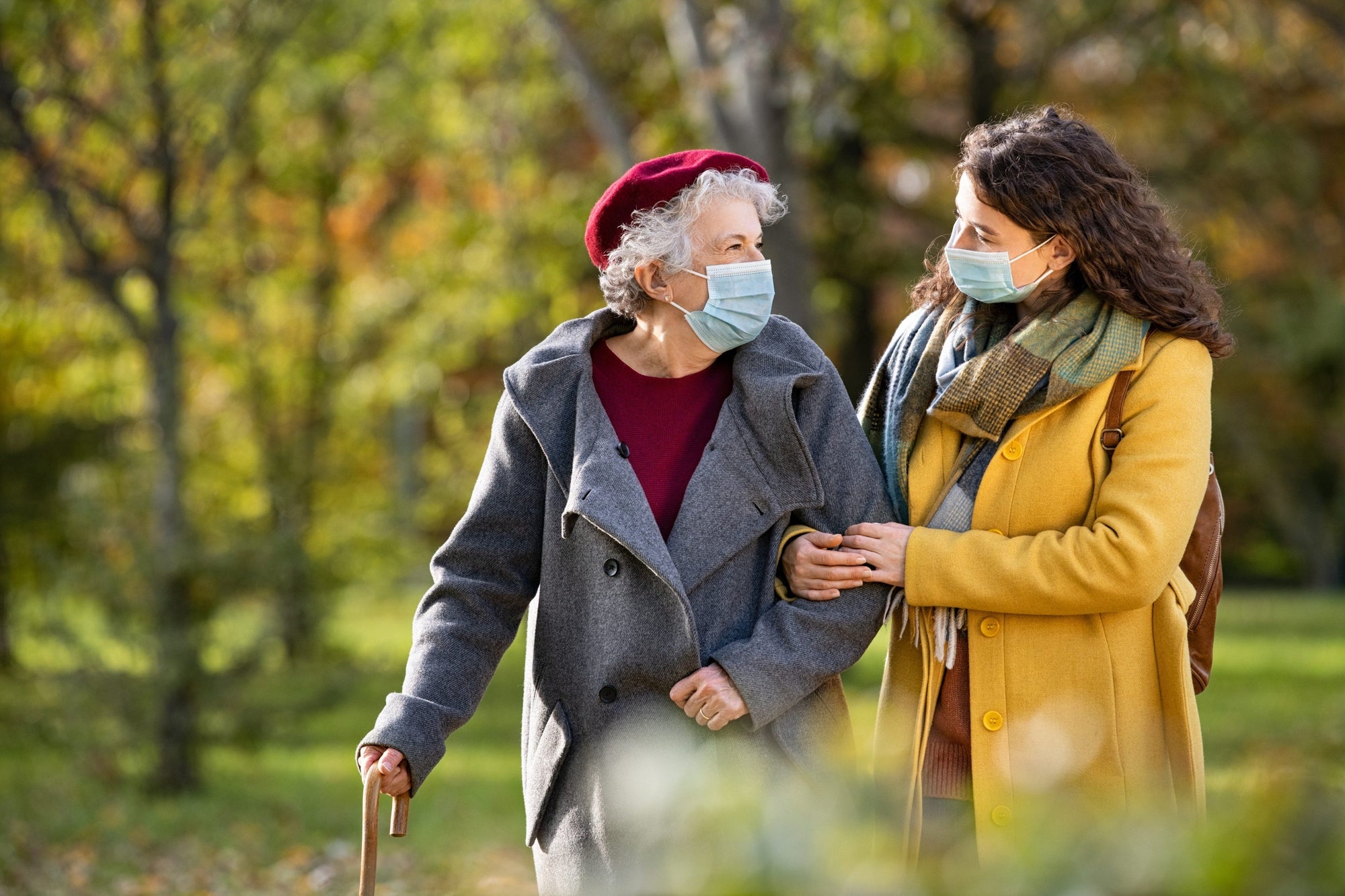 Daughter holds elderly mother, both wearing masks as they walk through a park