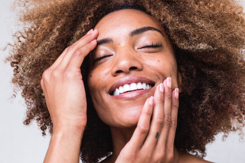 woman with darker complexion and glowing skin touching her face while smiling